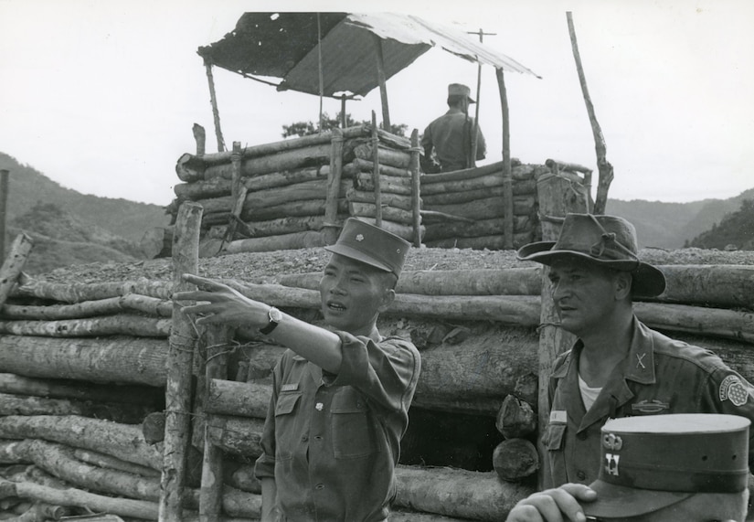 Two men look into the distance as one points. Behind them is a log fence and a taller lookout post also made of logs.