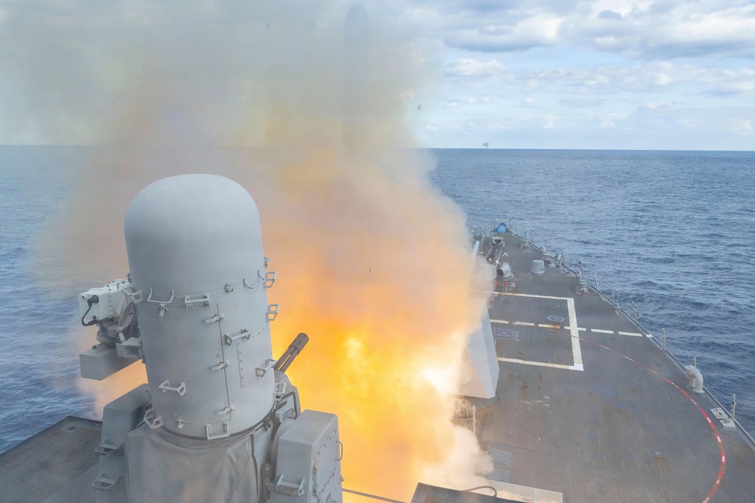 A ship at sea fires a weapon, creating a smoky fireball during the day.