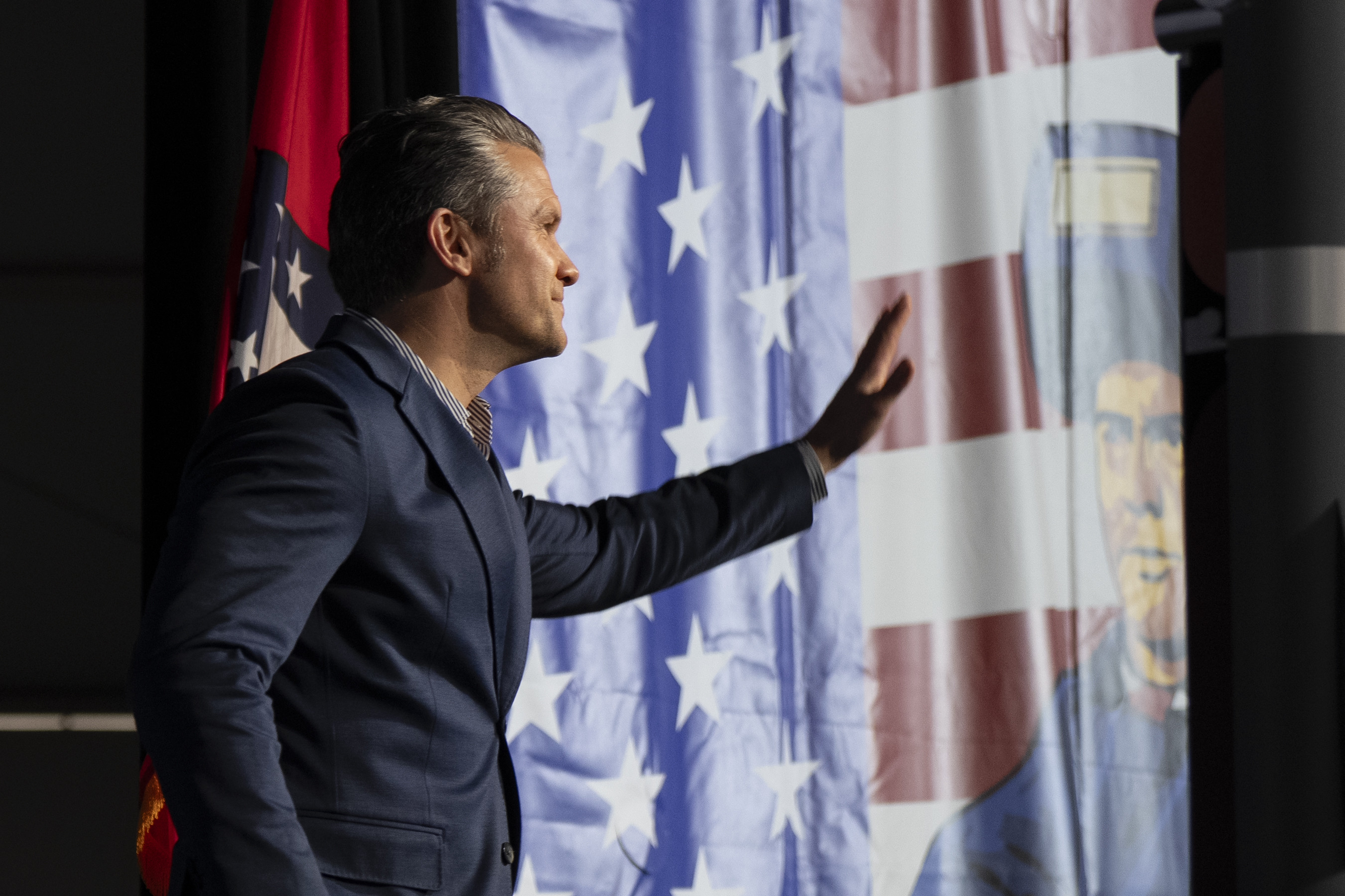 A person in a business suit stands in front of an American flag and waves with one hand.  