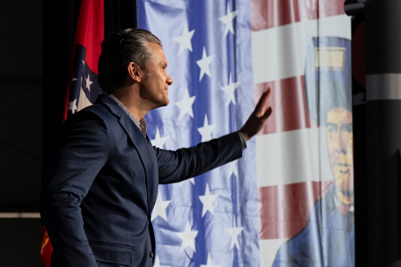A person in a business suit stands in front of an American flag and waves with one hand. 