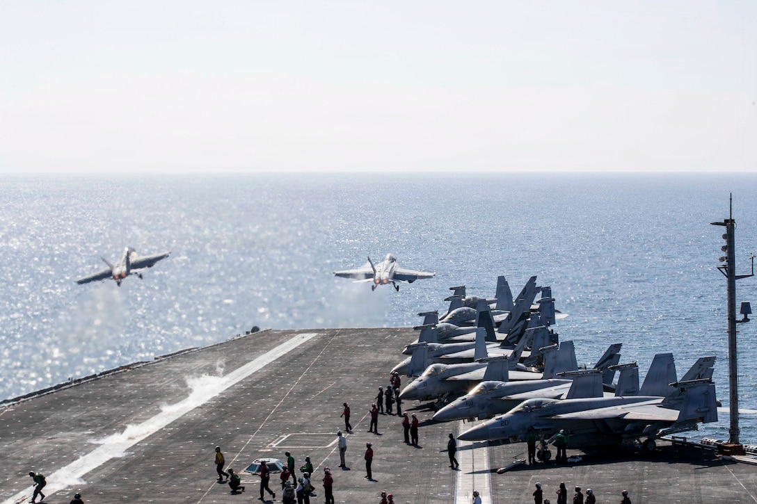 Two aircraft launch from the flight deck of a ship at sea during the day as dozens of fellow sailors gather nearby.