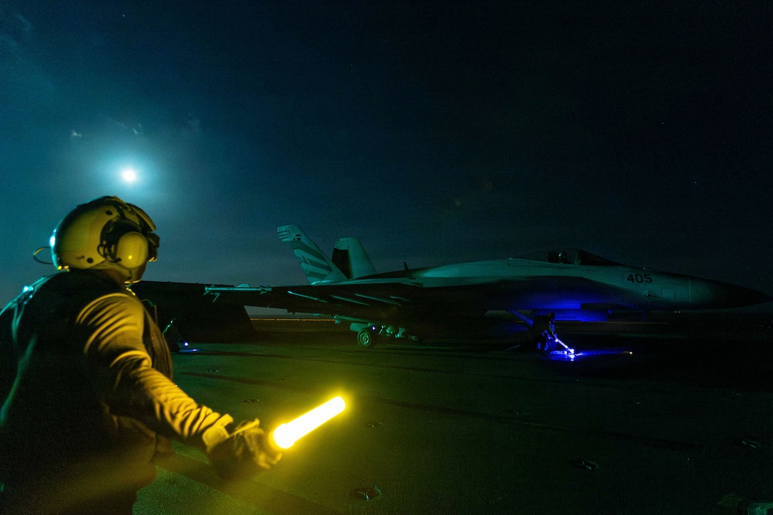 A sailor holding a yellow baton in the dark signals to an aircraft illuminated by a blue light on the flight deck of a ship.