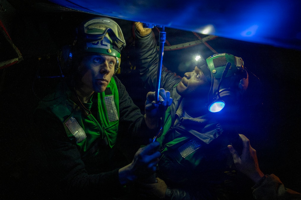 Sailors in green vests and helmets use flashlights and tools to repair a partially visible aircraft in the dark.