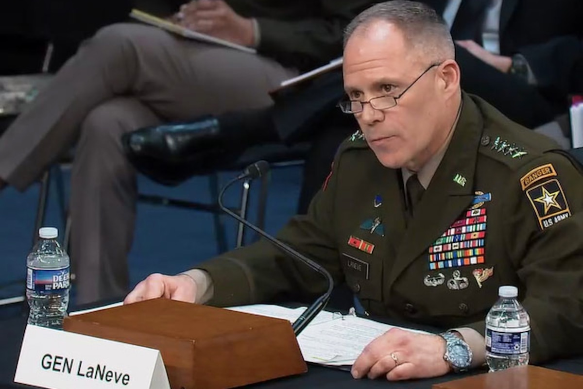 A man seated at a table wearing a formal military uniform speaks into a microphone.