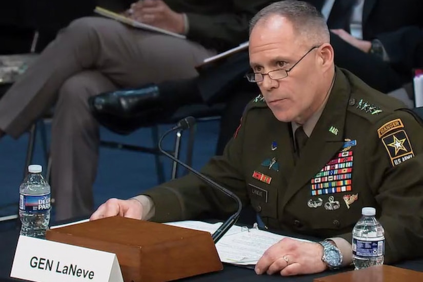 A man seated at a table wearing a formal military uniform speaks into a microphone.