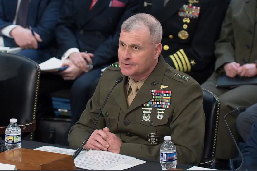 A man in a military dress uniform.is seated at a table speaking into a microphone.