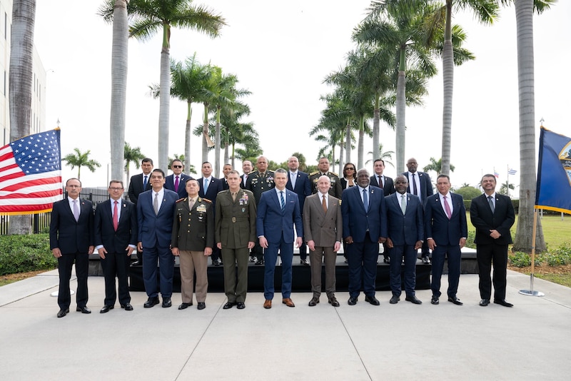 People dressed in business attire and formal military uniforms stand in two rows outside. Behind them are palm trees, and on either side are an American flag and a blue flag.