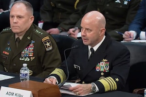 A man in a military dress uniform is seated at a table speaking into a microphone. There is another man in similar attire seated next to him.