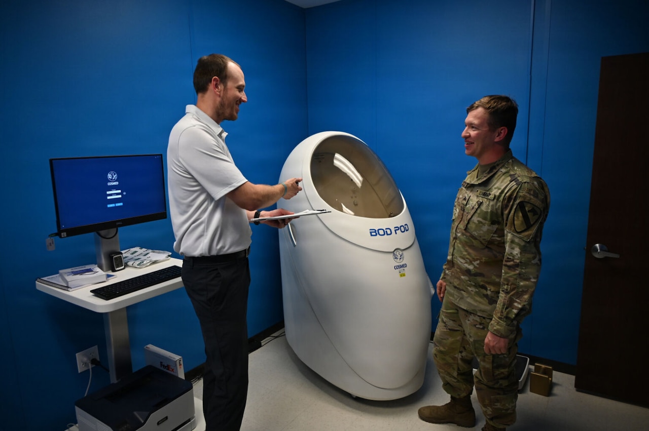 A person in business attire gestures toward a white capsule while talking to a man in a camouflage military uniform.