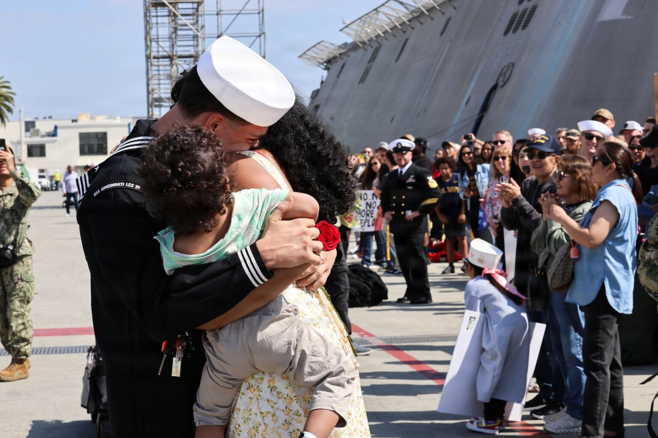 A man in a military dress uniform hugs a woman and child in front of a crowd on a dock. There is a large military ship on the right.
