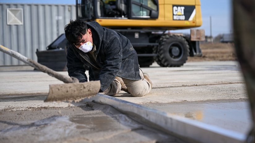A person uses a long metal plank to smooth the top of poured concrete while another person kneels on the ground leveling it out.