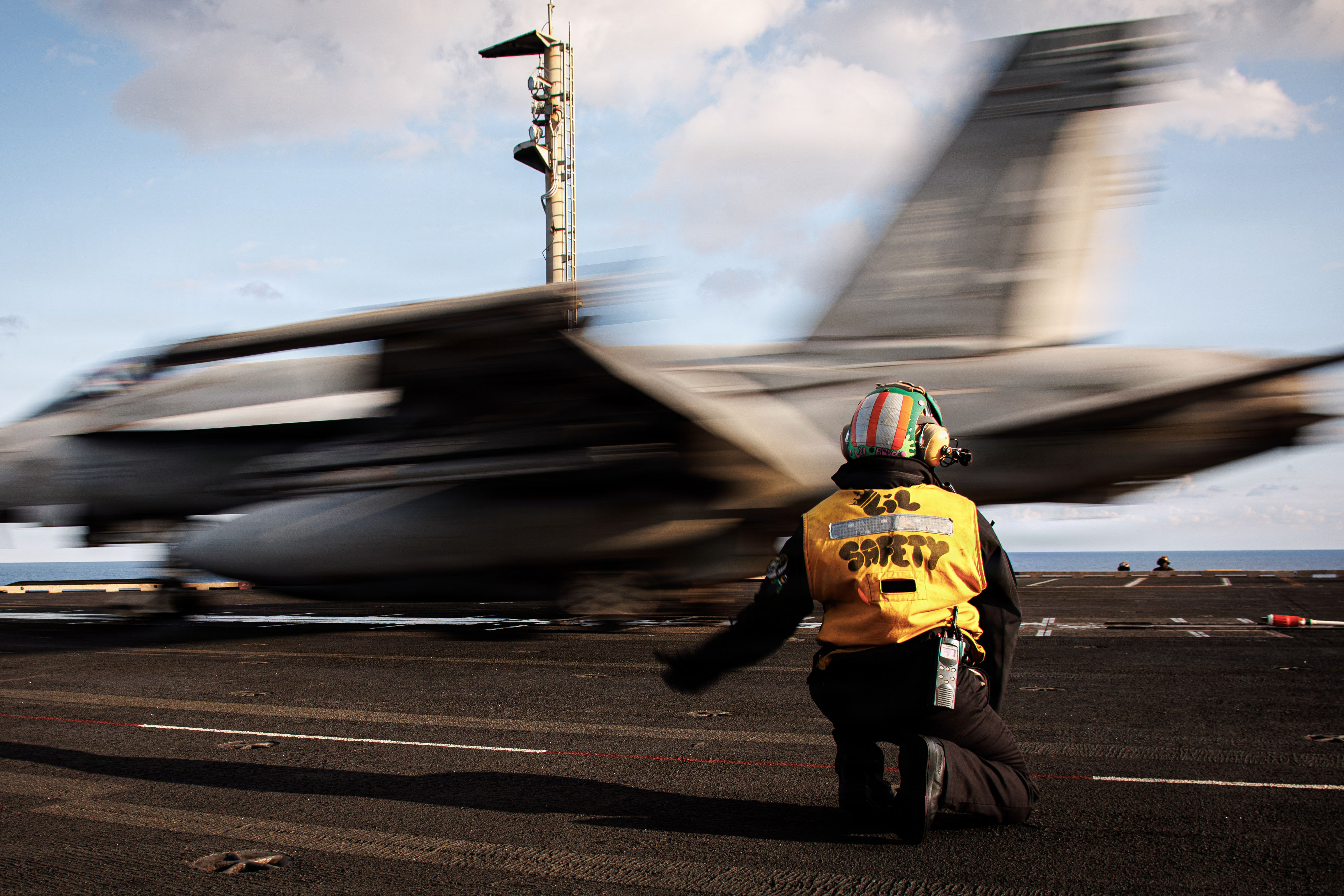 A Navy jet speeds by a sailor in a yellow vest, shown from behind, signaling on a ship's flight dec..
