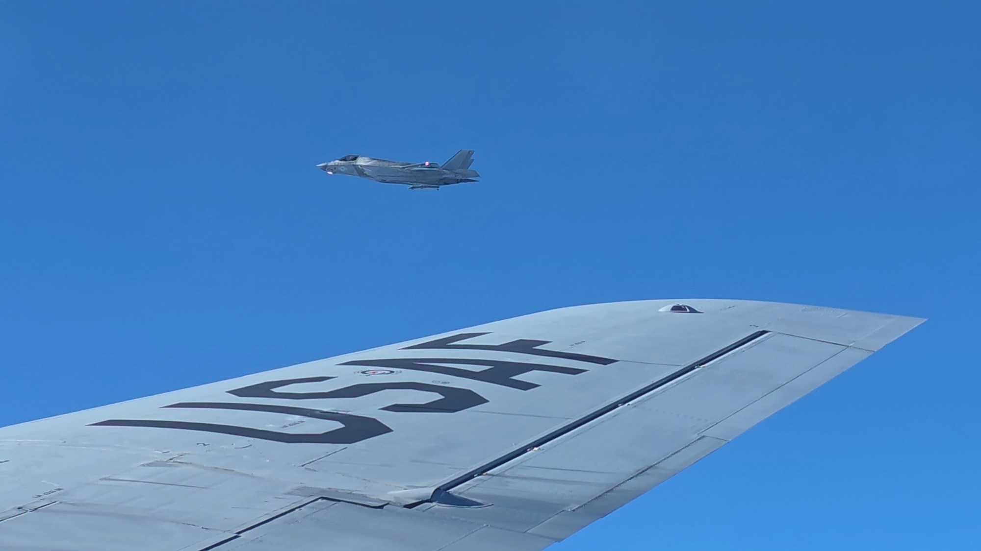 A blue sky, with a jet towards the center of the photo. From the bottom to the center, there is a wing of another plane. The planes are flying next to each other.