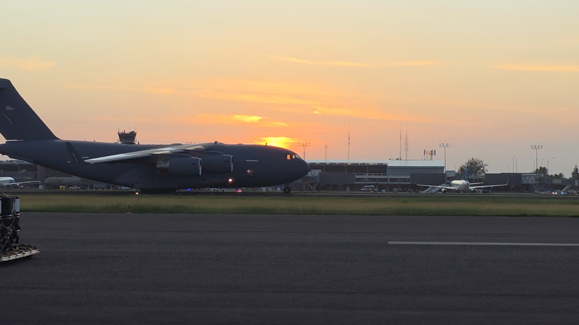 Orange hues fill the sky, with a large plane in the center of the photo. The plane is located on a flightline.