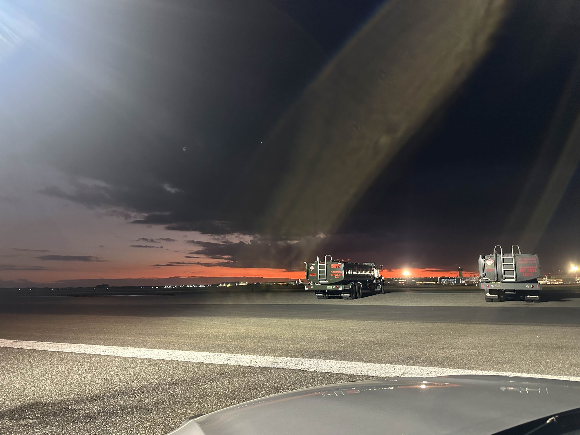 A dark, but lit, flightline. There are refueler trucks on the strip, with an orange hue along the skyline.