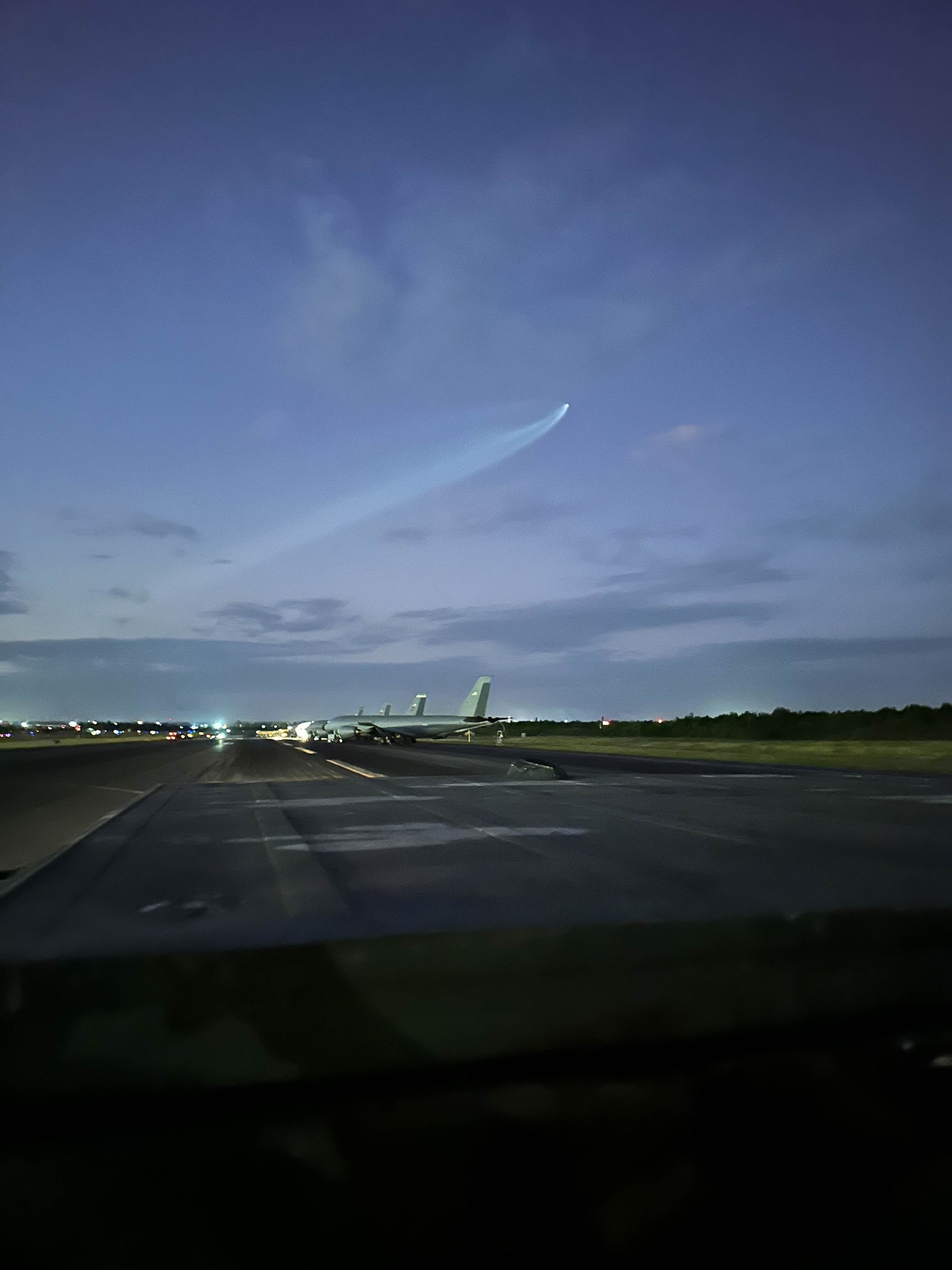 A dark photo of a long strip of road. There are planes in the distance, with another plane taking flight higher up in the sky.