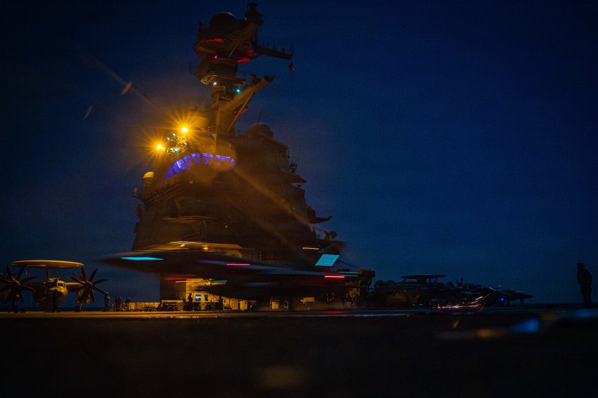 An aircraft flies past the mast of a ship as it prepares to land on the deck at night.