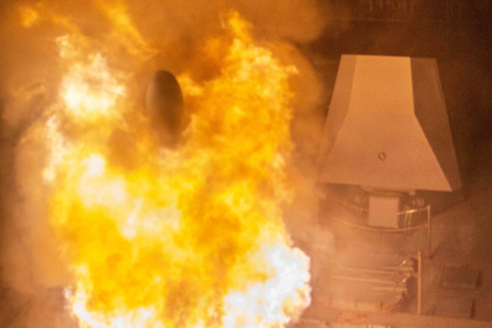 Flames erupt from a ship's missile hatch at night. At the center of the flames is the tip of a just-launched missile.