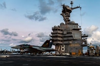 A fighter jet touches down on the flight deck of an aircraft carrier; the ship's tall, multileveled superstructure is in the background.