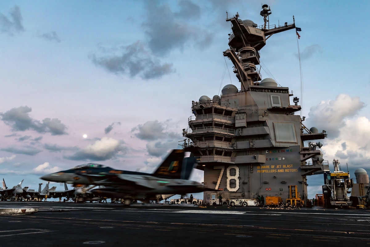 A fighter jet touches down on the flight deck of an aircraft carrier; the ship's tall, multileveled superstructure is in the background.