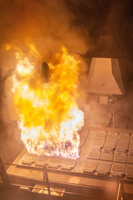 Flames erupt from a ship's missile hatch at night. At the center of the flames is the tip of a just-launched missile.