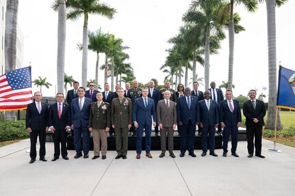 Nearly two dozen men and one woman dressed in business attire and formal military uniforms stand in two lines outside. Behind them are two rows of palm trees, and on either side are an American flag and a blue flag.