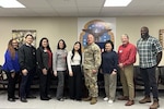 A group of nine people, one wearing a camouflage military uniform, pose for a group photo indoors in a conference room.