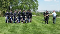 On the left is a group of Army Soldiers in ceremonial uniforms marching in a circle with rifles in their hands as a video crew of two members, on the right, are filming their actions while on a bright green lawn that overlooks Washington, D.C.