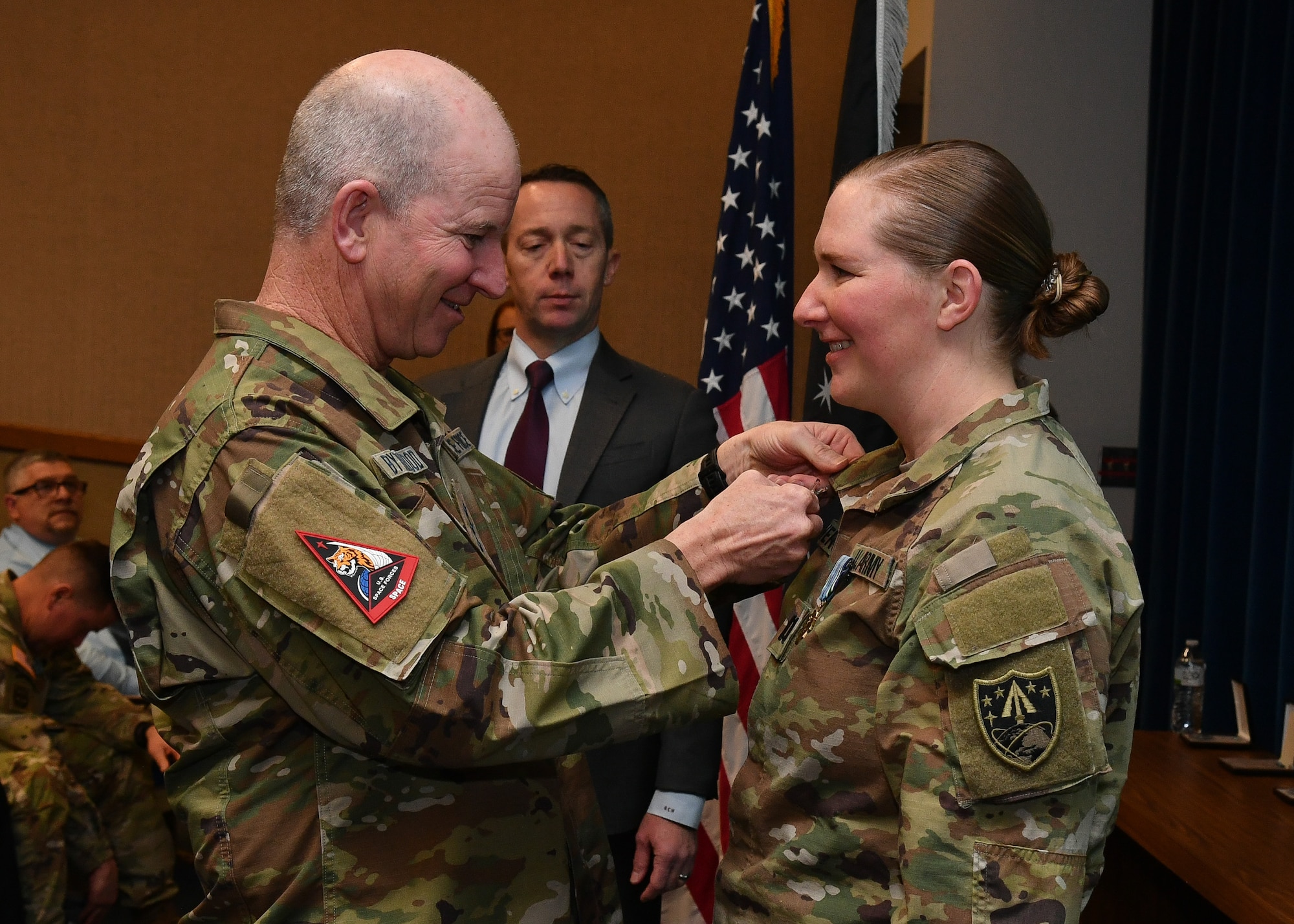 A military man pins affixes a medal to the collar of a military woman in an auditorium while both smile