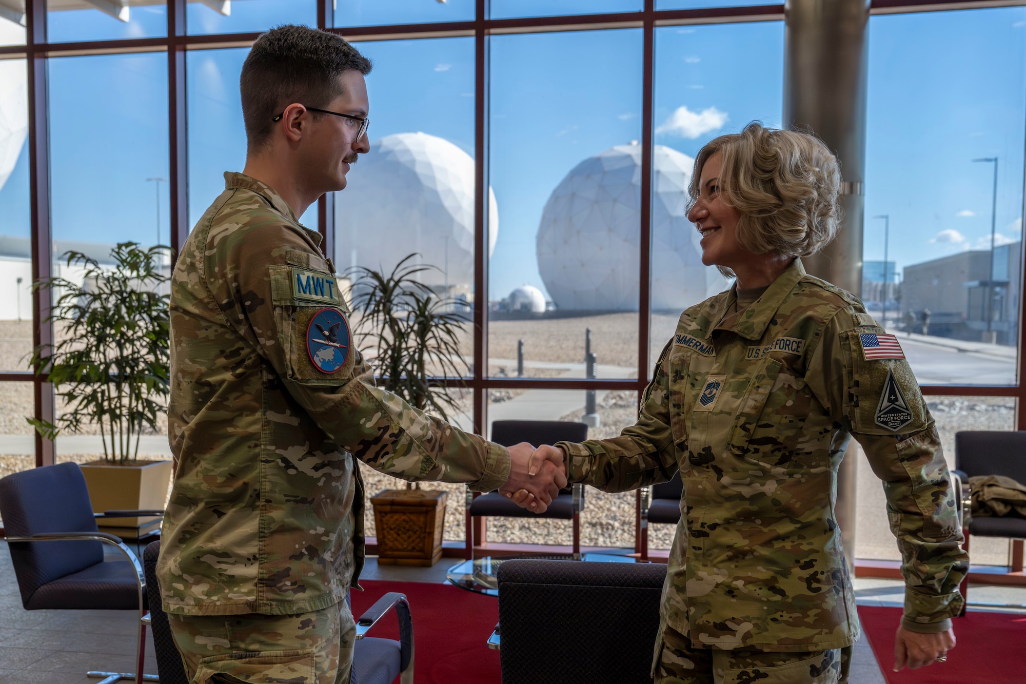 A military woman shakes hands with a military man in front of a window with a view of two giant space sensors