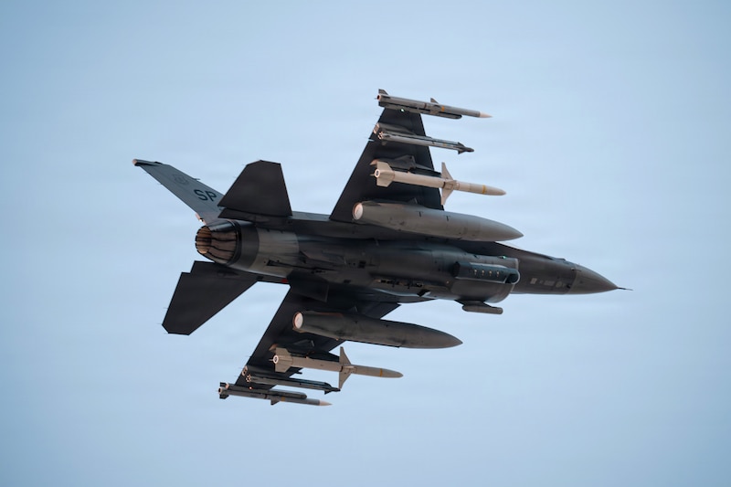 An aircraft carrying ordnance flies in a blue sky as seen from below.