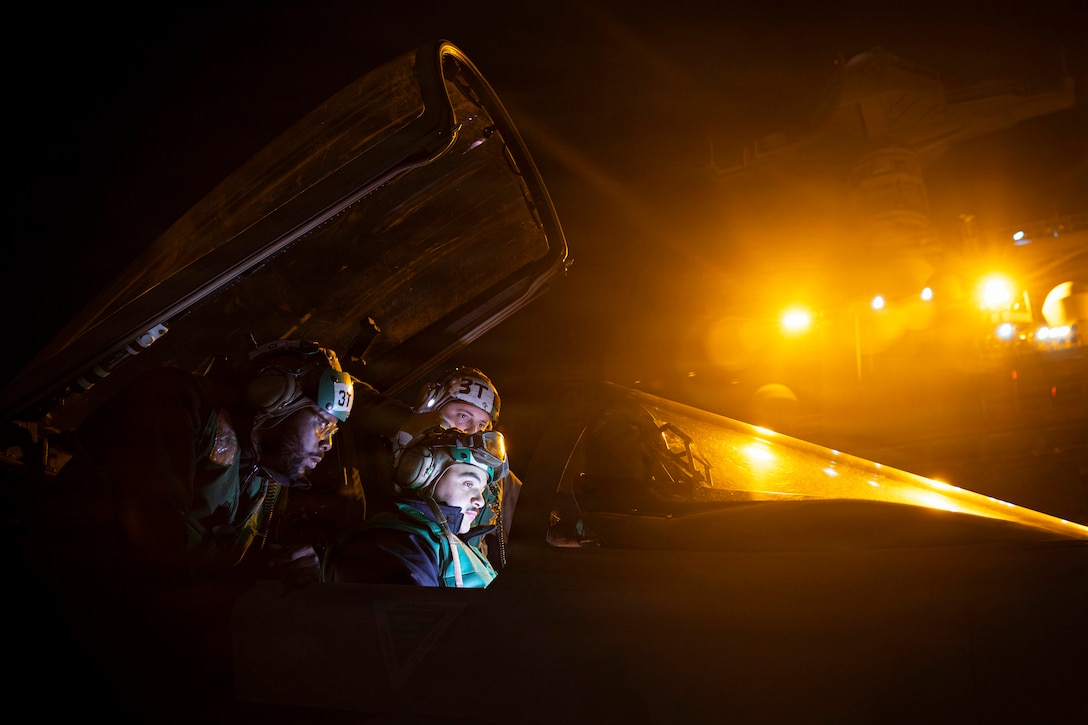 Three sailors wearing helmets sit in the open cockpit of an aircraft on a flight deck illuminated by yellowish ship lights from above.