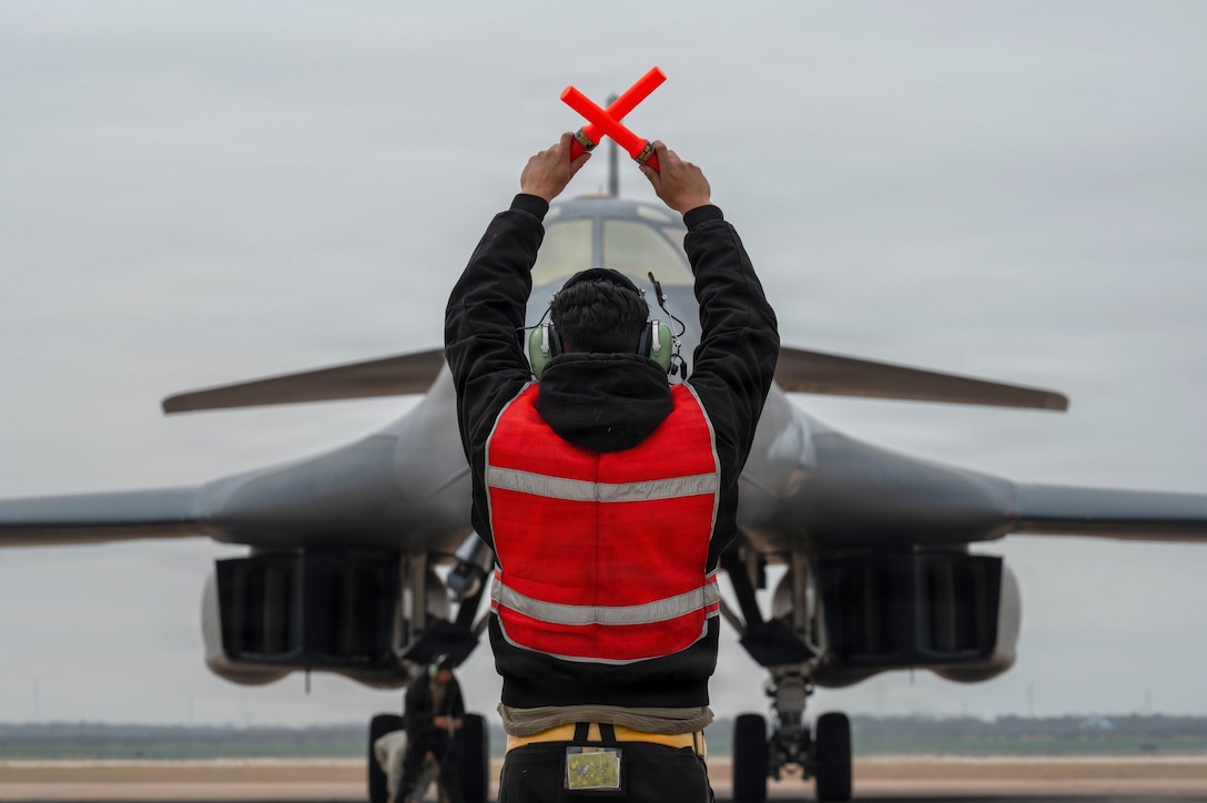An airman signaling with orange batons stands in front of an aircraft on a tarmac on a gloomy day.