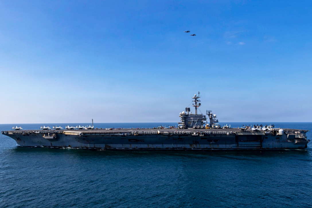 Two aircraft fly in formation in a blue sky over a naval ship in a body of water.
