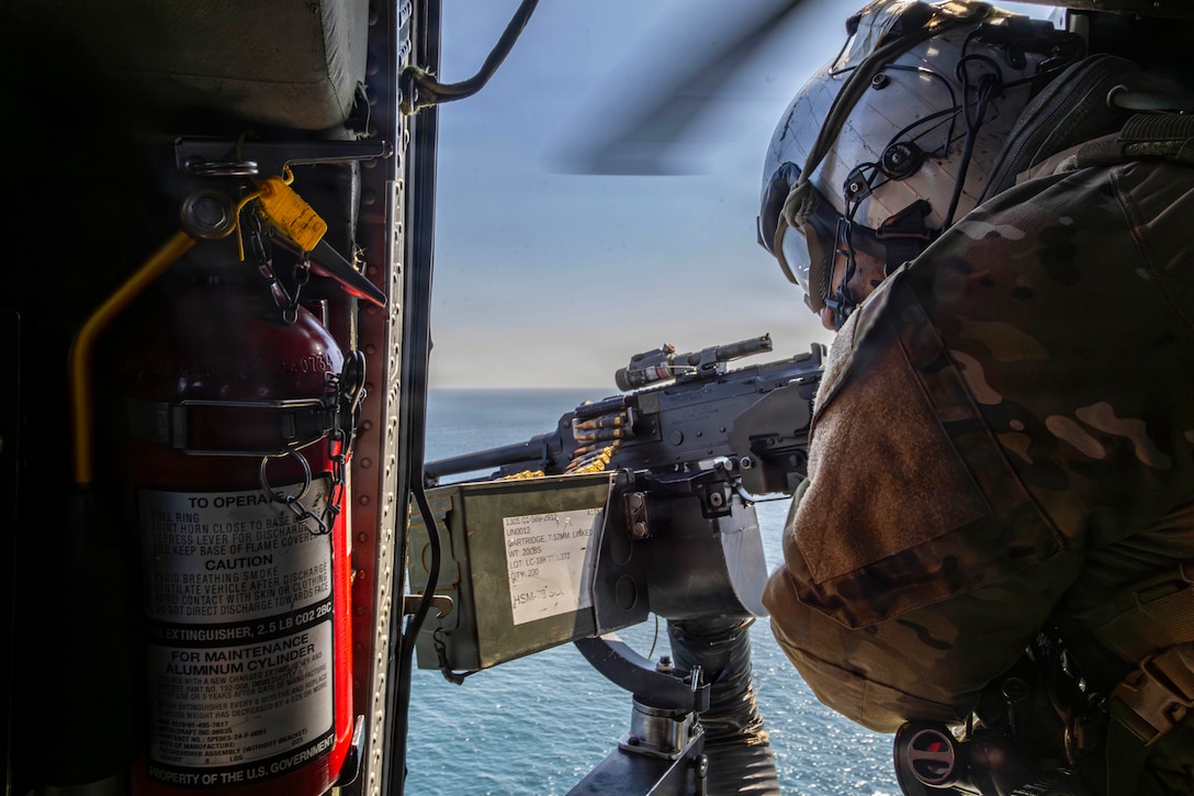 A sailor aims a weapon toward blue waters out of the open doorway of an airborne helicopter.