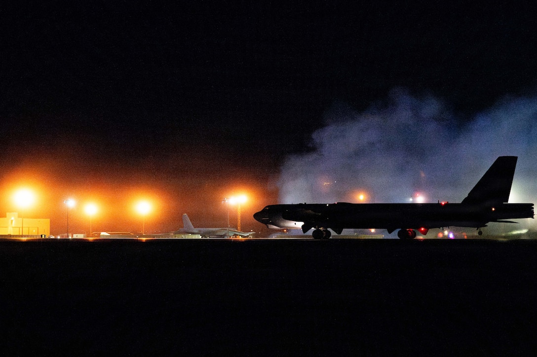 An aircraft sits next to a cloud of smoke in the dark on a tarmac illuminated by yellowish streetlights.