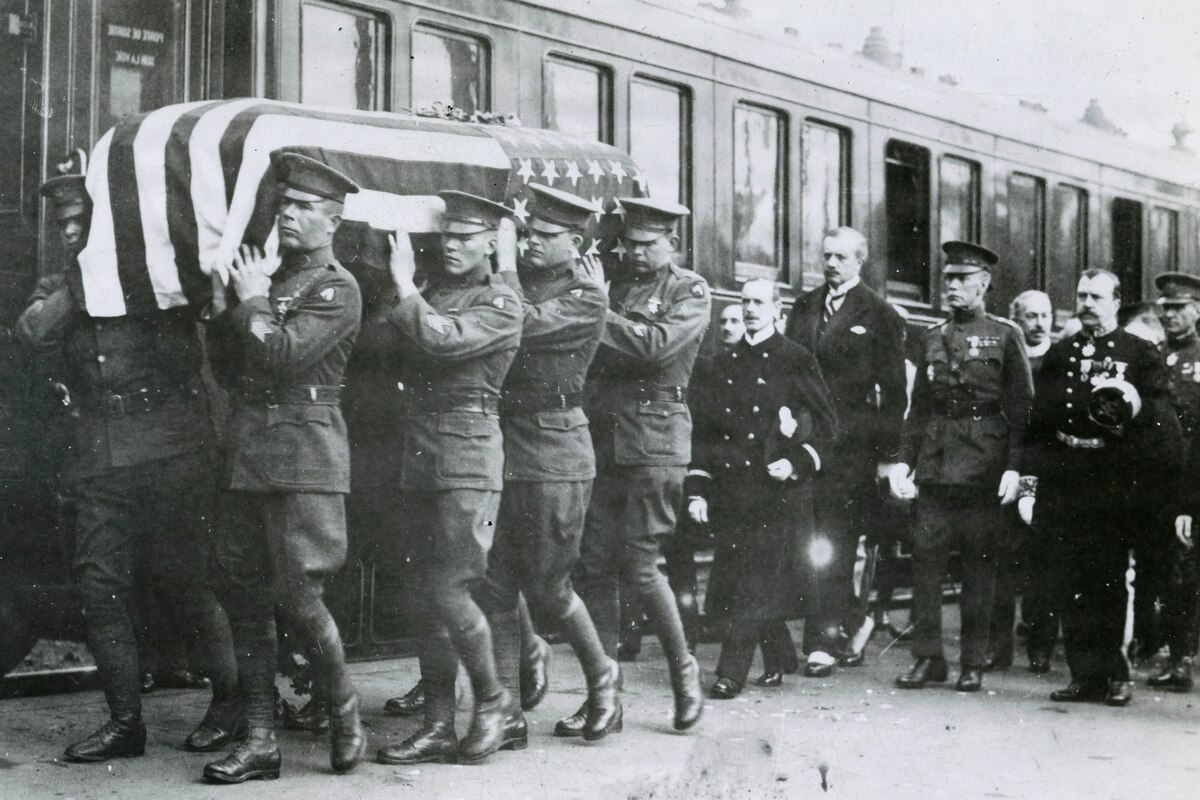 In a black and white photo, men in military dress uniforms carry a casket draped in the American flag next to a train. Six men in military dress uniforms walk behind the casket.