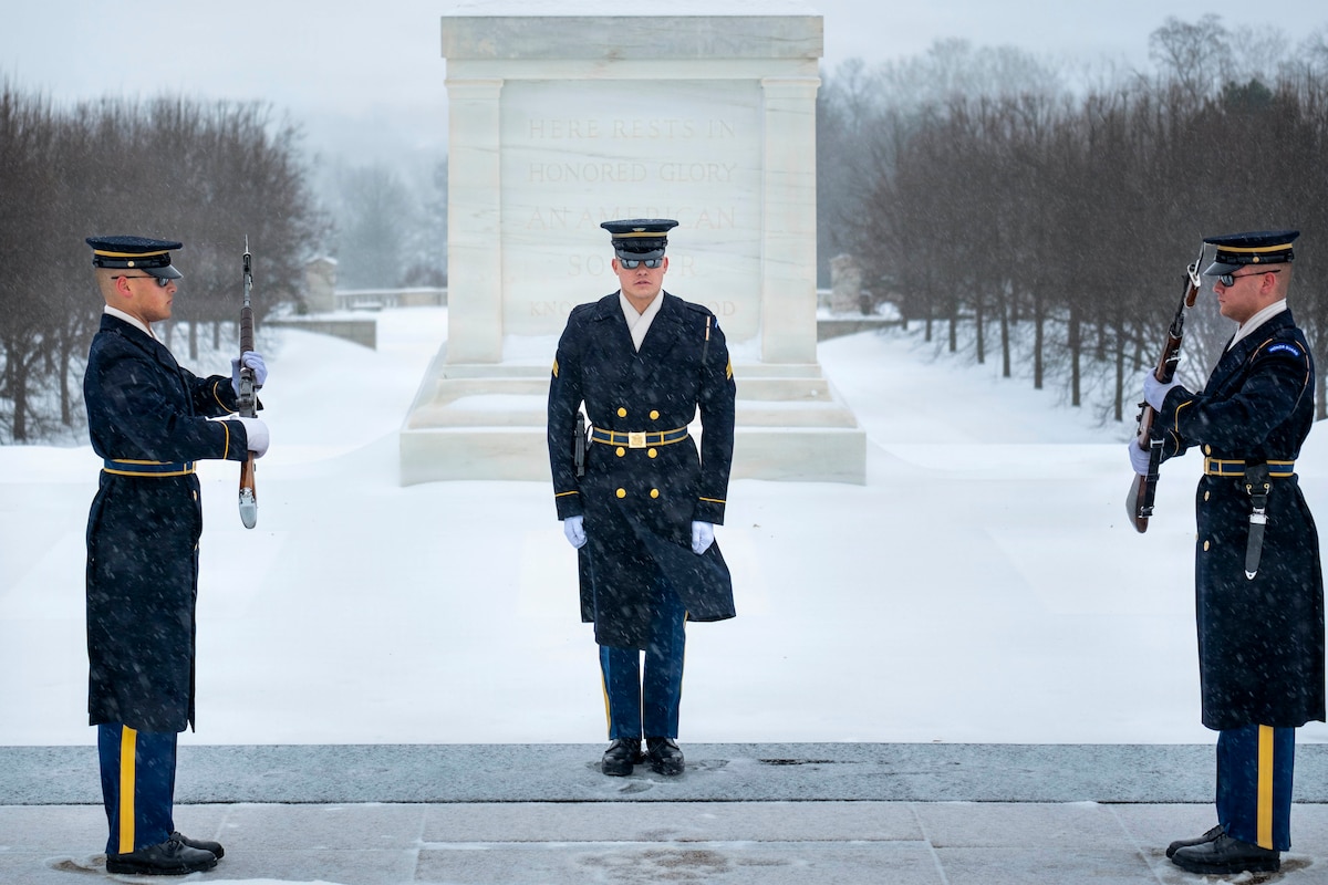 Standing on a snowy platform outside, two men in military dress uniforms face each other, holding rifles in their hands, as another man in similar attire observes. There is a snow-covered field and a memorial in the background.