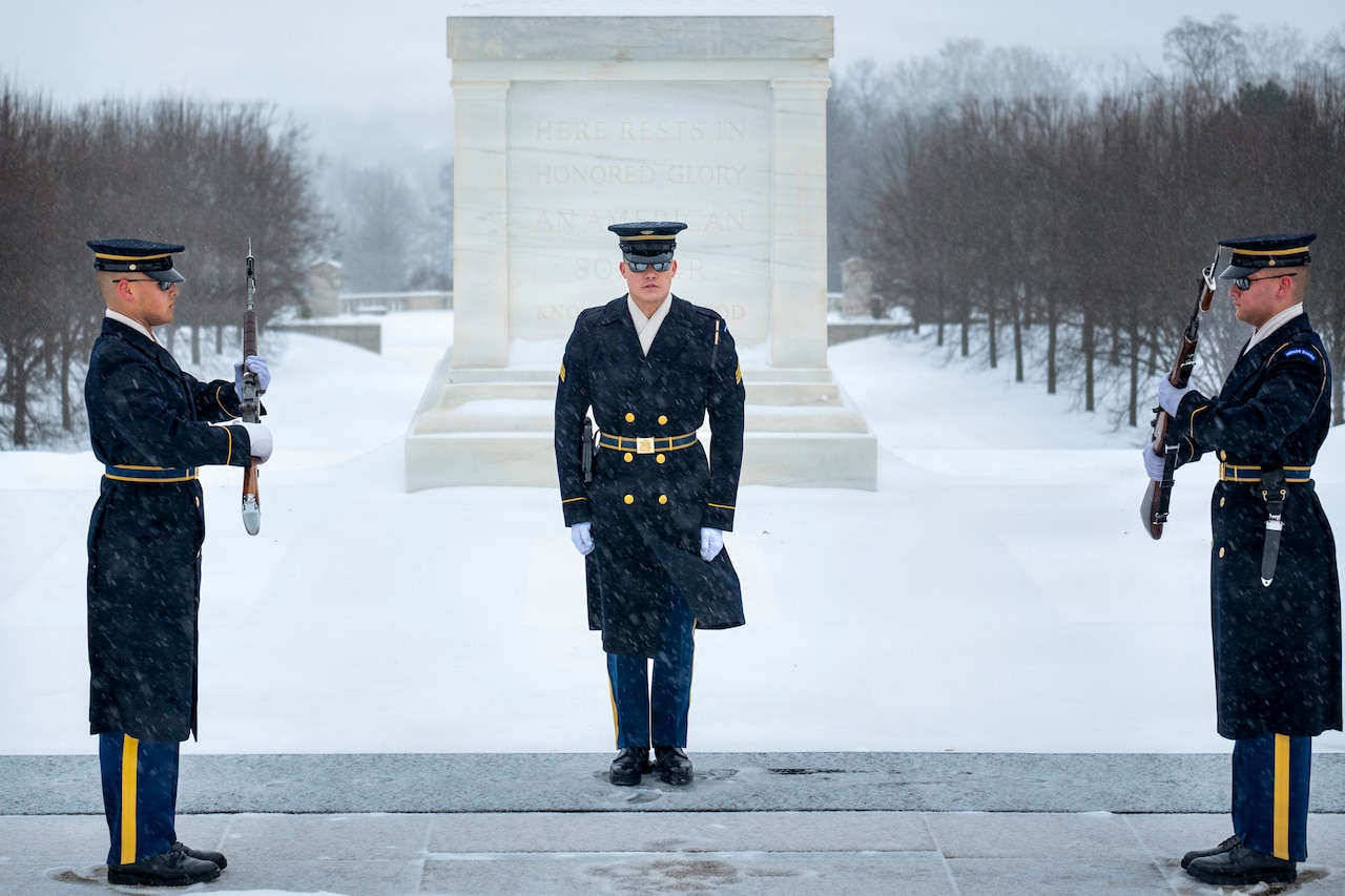 Standing on a snowy platform outside, two men in military dress uniforms face each other, holding rifles in their hands, as another man in similar attire observes. There is a snow-covered field and a memorial in the background.