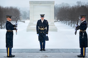 Standing on a snowy platform outside, two men in military dress uniforms face each other, holding rifles in their hands, as another man in similar attire observes. There is a snow-covered field and a memorial in the background.