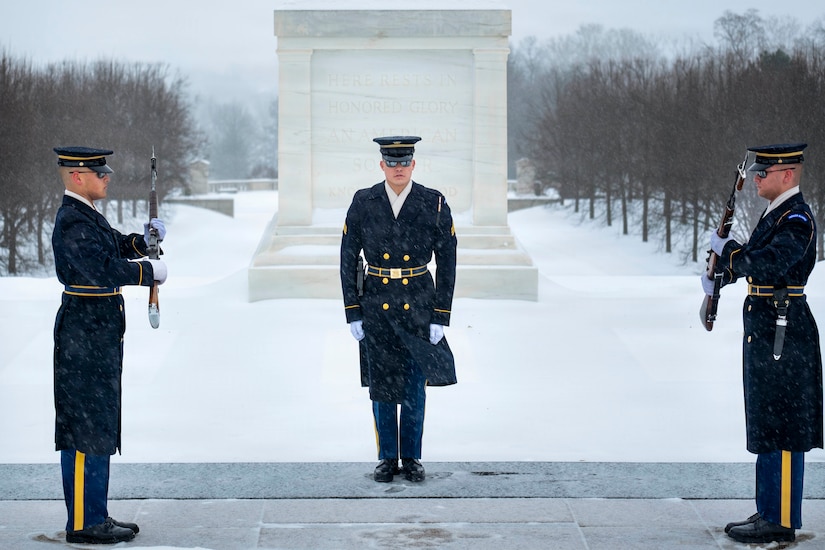 Standing on a snowy platform outside, two men in military dress uniforms face each other, holding rifles in their hands, as another man in similar attire observes. There is a snow-covered field and a memorial in the background.