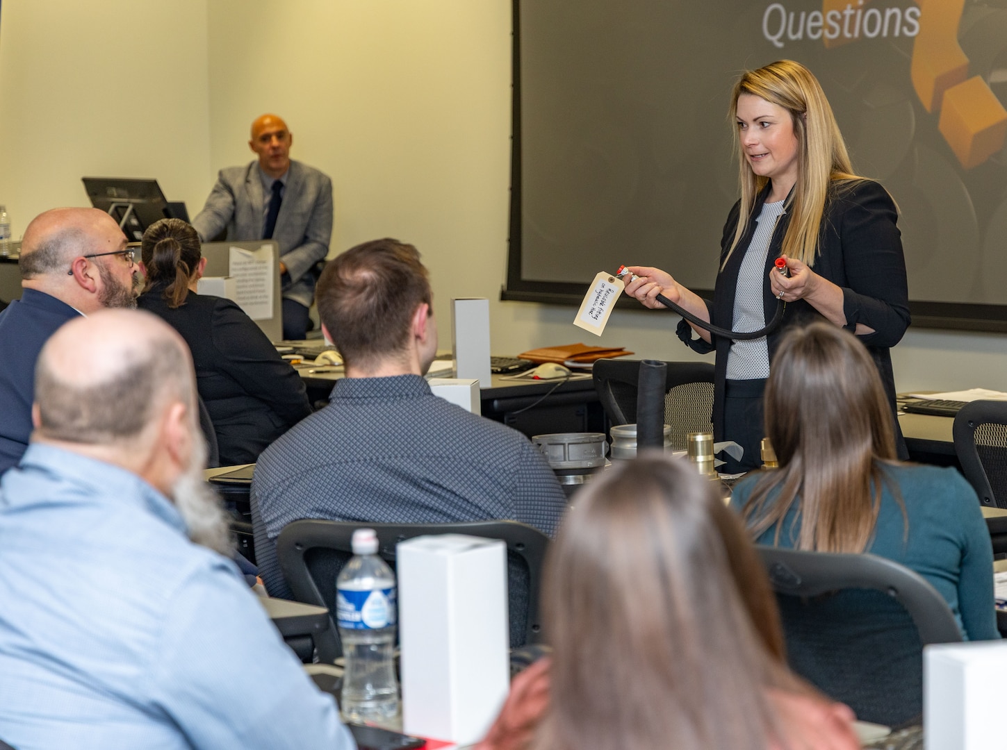 A light-haired woman shows a hose to a group of people in a classroom.