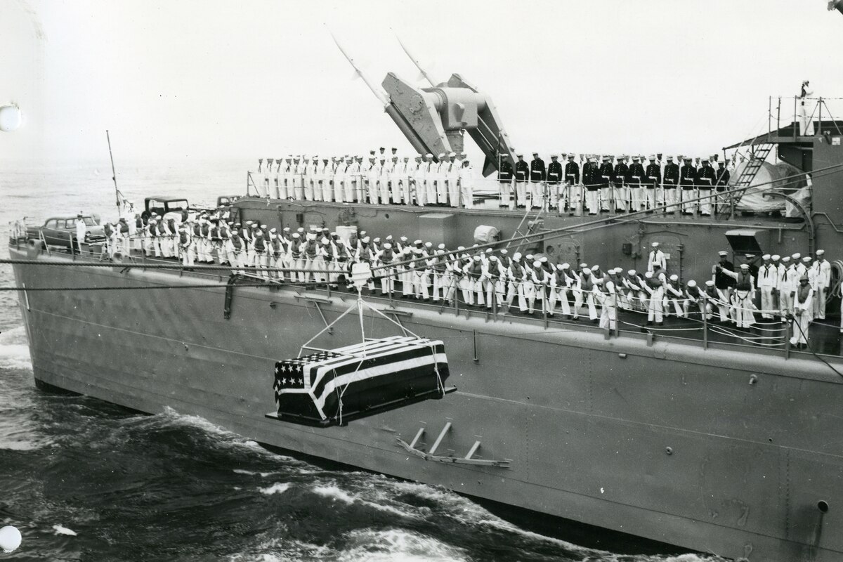 In a black and white photo, a casket draped in the American flag hangs from a rope as it travels from one ship to another. There is a large military ship in the background with dozens of people standing on the deck in military dress uniforms.