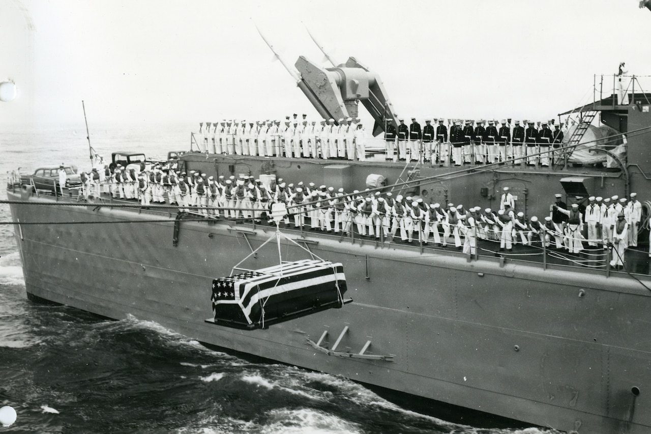 In a black and white photo, a casket draped in the American flag hangs from a rope as it travels from one ship to another. There is a large military ship in the background with dozens of people standing on the deck in military dress uniforms.