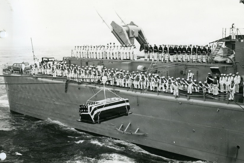 In a black and white photo, a casket draped in the American flag hangs from a rope as it travels from one ship to another. There is a large military ship in the background with dozens of people standing on the deck in military dress uniforms.