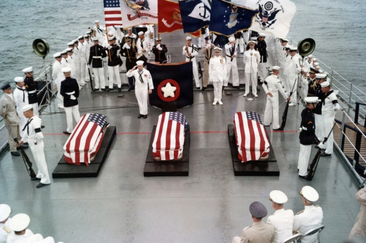 On the deck of a large military ship, dozens of people in military dress uniforms stand, honoring three caskets draped in the American flag. The American flag, Navy flag and Marine Corps flag are being held in the background.
