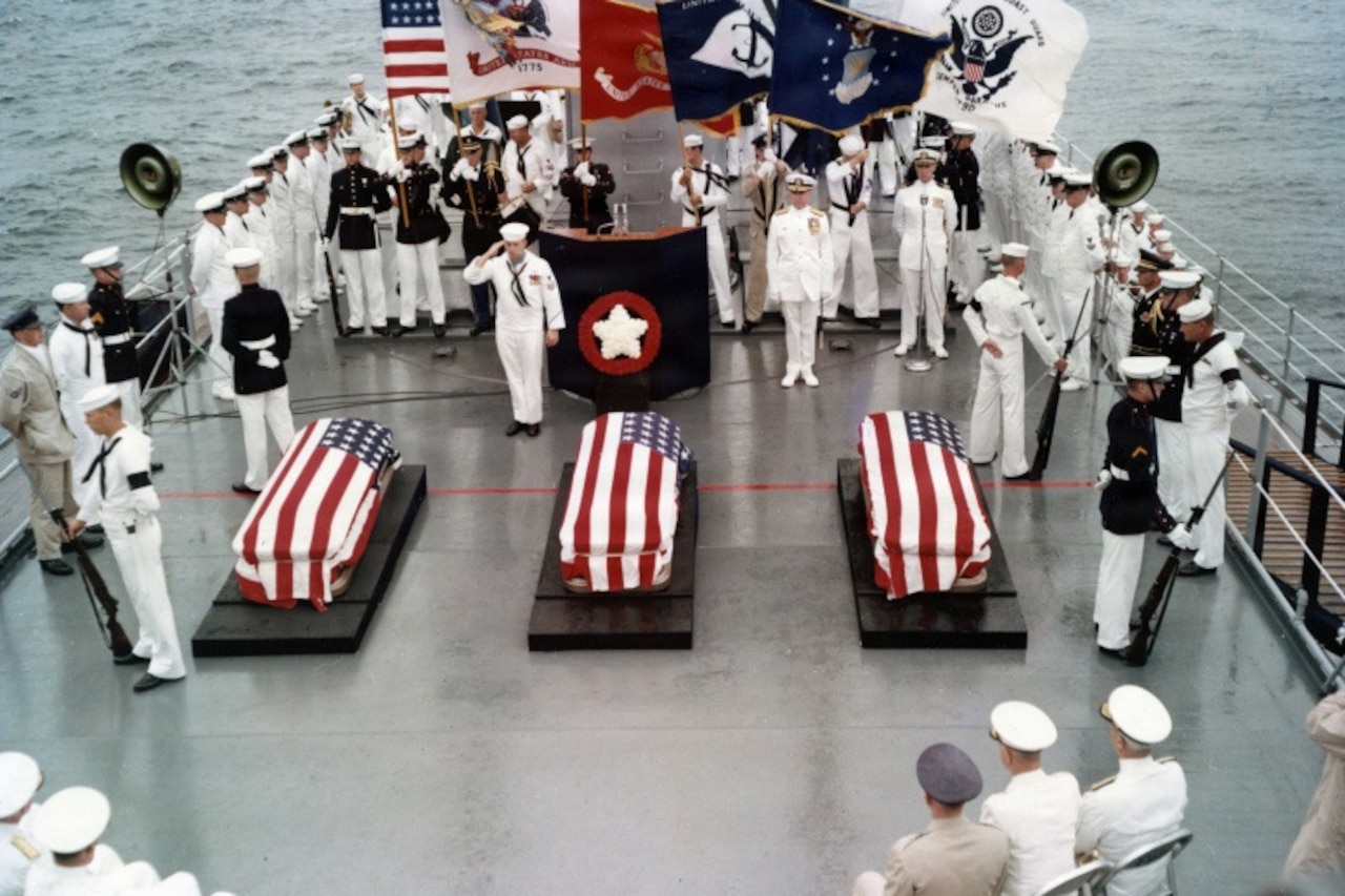 On the deck of a large military ship, dozens of people in military dress uniforms stand, honoring three caskets draped in the American flag. The American flag, Navy flag and Marine Corps flag are being held in the background.