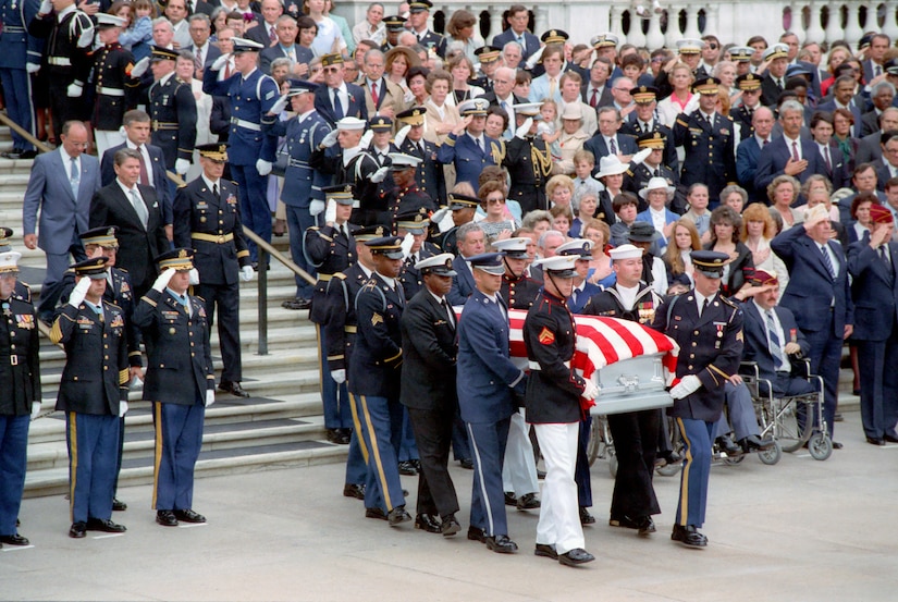 Men in military dress uniforms carry a casket draped in the American flag. Dozens of people in military dress uniforms and formal attire stand in rows on stairs as they salute the casket.