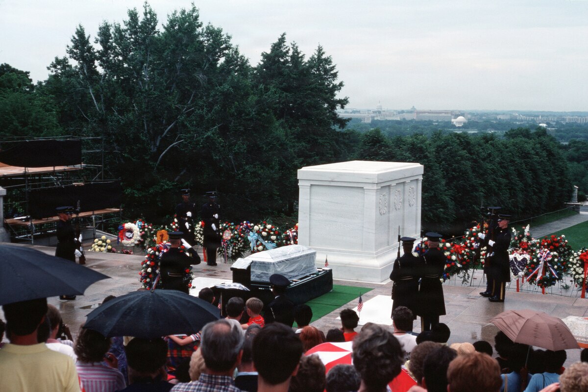 Men in military dress uniforms stand around a casket that is in front of a memorial. Dozens of people in casual attire stand in rows looking at the casket, some holding umbrellas.