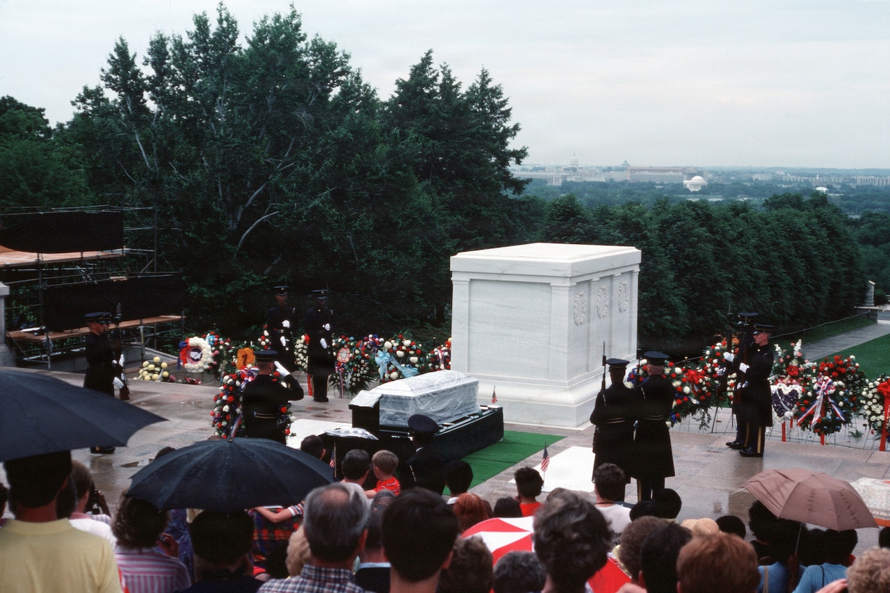 Men in military dress uniforms stand around a casket that is in front of a memorial. Dozens of people in casual attire stand in rows looking at the casket, some holding umbrellas.